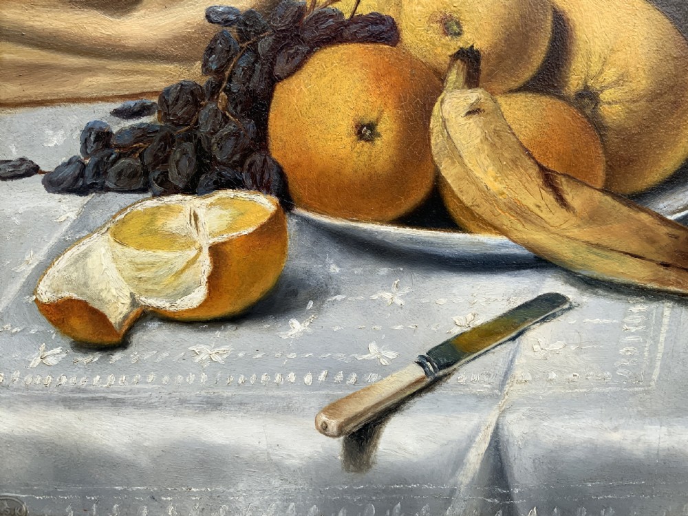 An Edwardian still life oil painting depicting citrus fruit, bananas, grapes, and a glass of wine on a tablecloth, set inside a heavy gold gilded frame. View IX