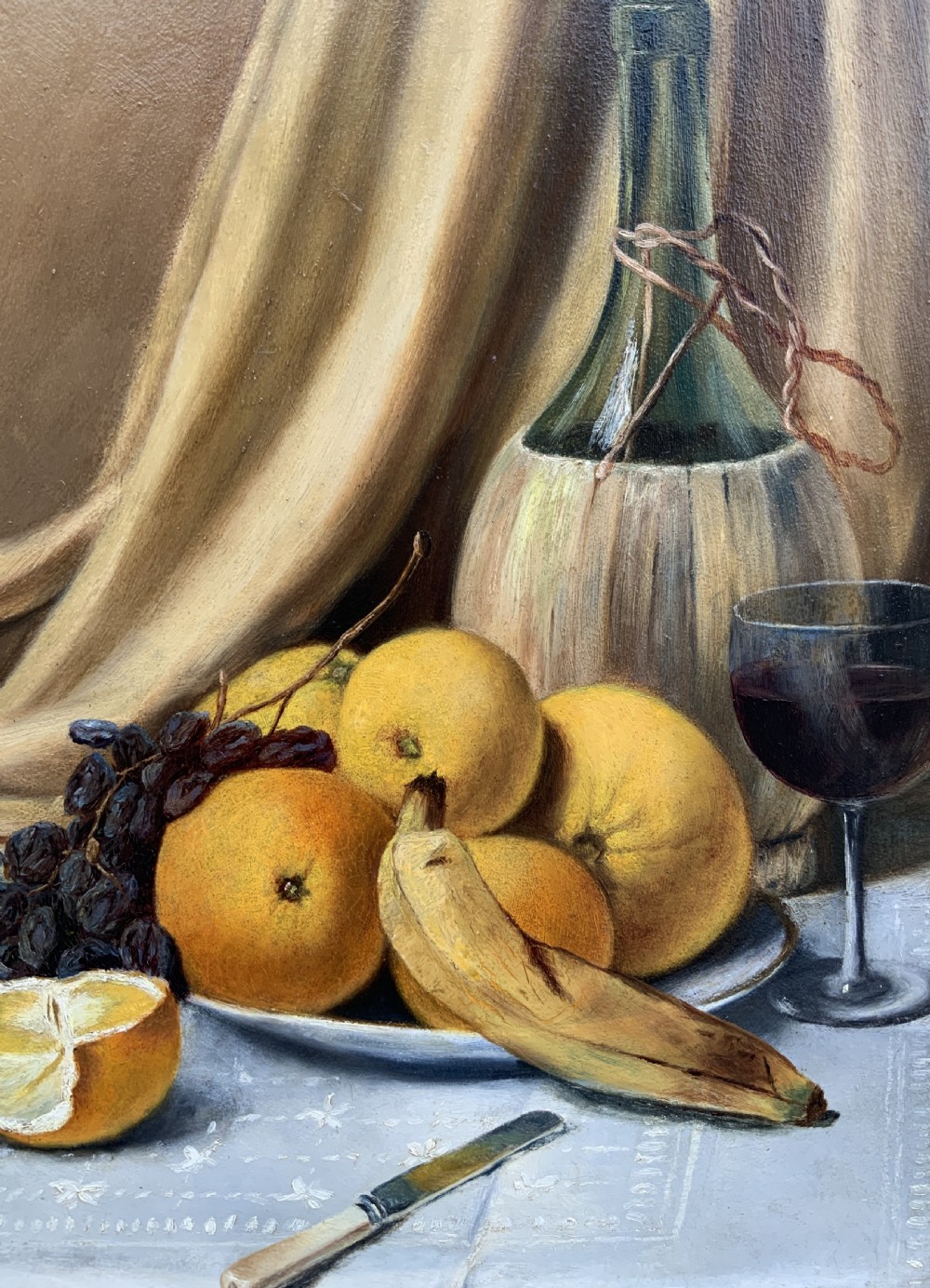 An Edwardian still life oil painting depicting citrus fruit, bananas, grapes, and a glass of wine on a tablecloth, set inside a heavy gold gilded frame. View X