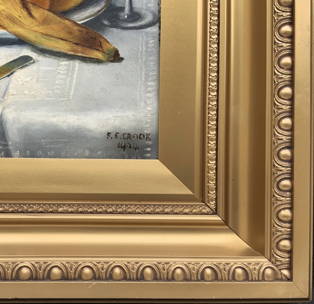 An Edwardian still life oil painting depicting citrus fruit, bananas, grapes, and a glass of wine on a tablecloth, set inside a heavy gold gilded frame. View XI