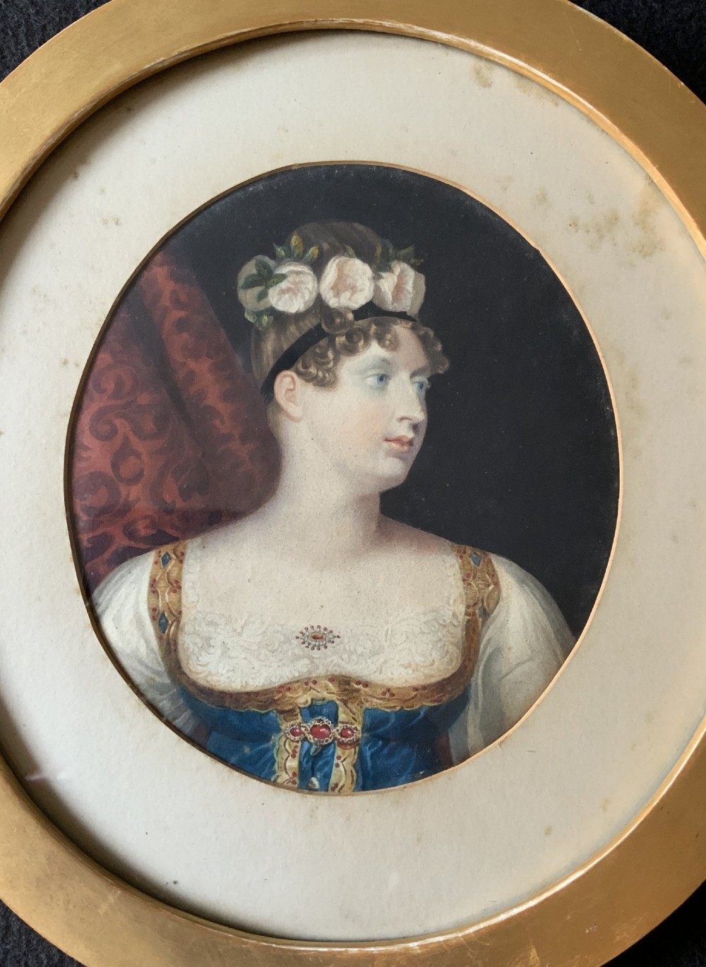 A circular gilt-framed hand-coloured miniature portrait depicting a young woman wearing a floral headband and blue dress. View III