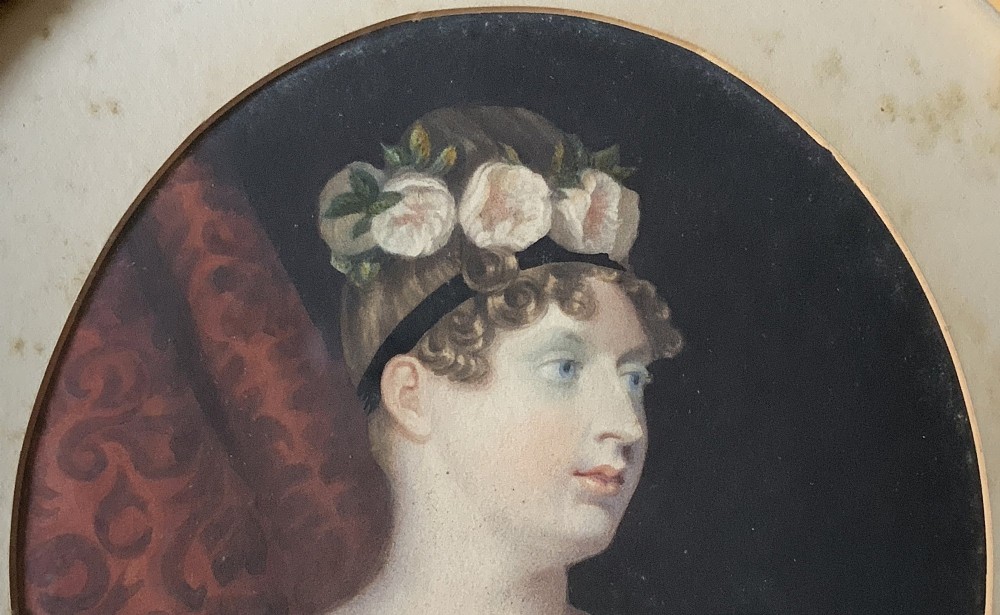 A circular gilt-framed hand-coloured miniature portrait depicting a young woman wearing a floral headband and blue dress. View V