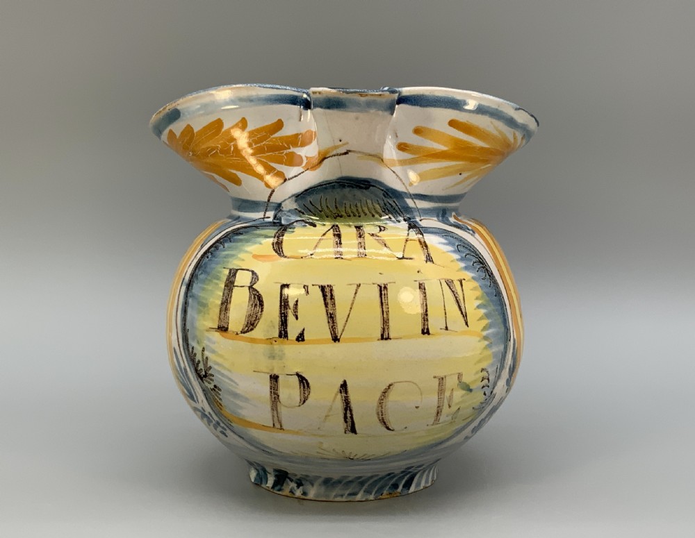 An antique Italian majolica earthenware pharmacy jug with hand-painted yellow, blue, and manganese script reading CARA BEVI IN PACE.