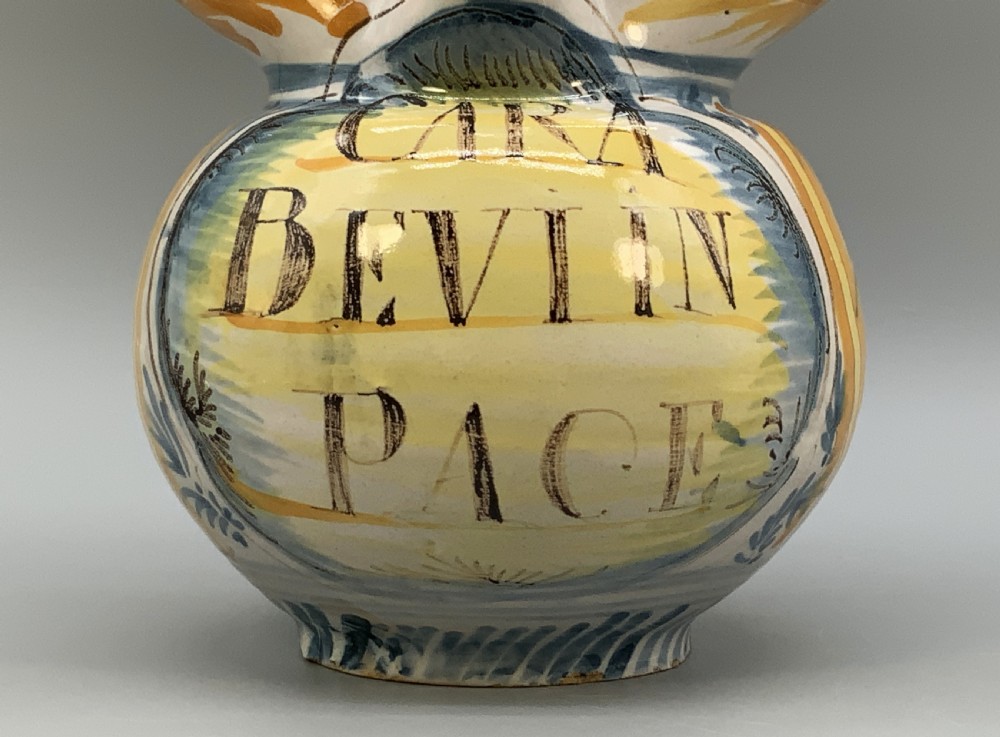 An antique Italian majolica earthenware pharmacy jug with hand-painted yellow, blue, and manganese script reading CARA BEVI IN PACE. View X