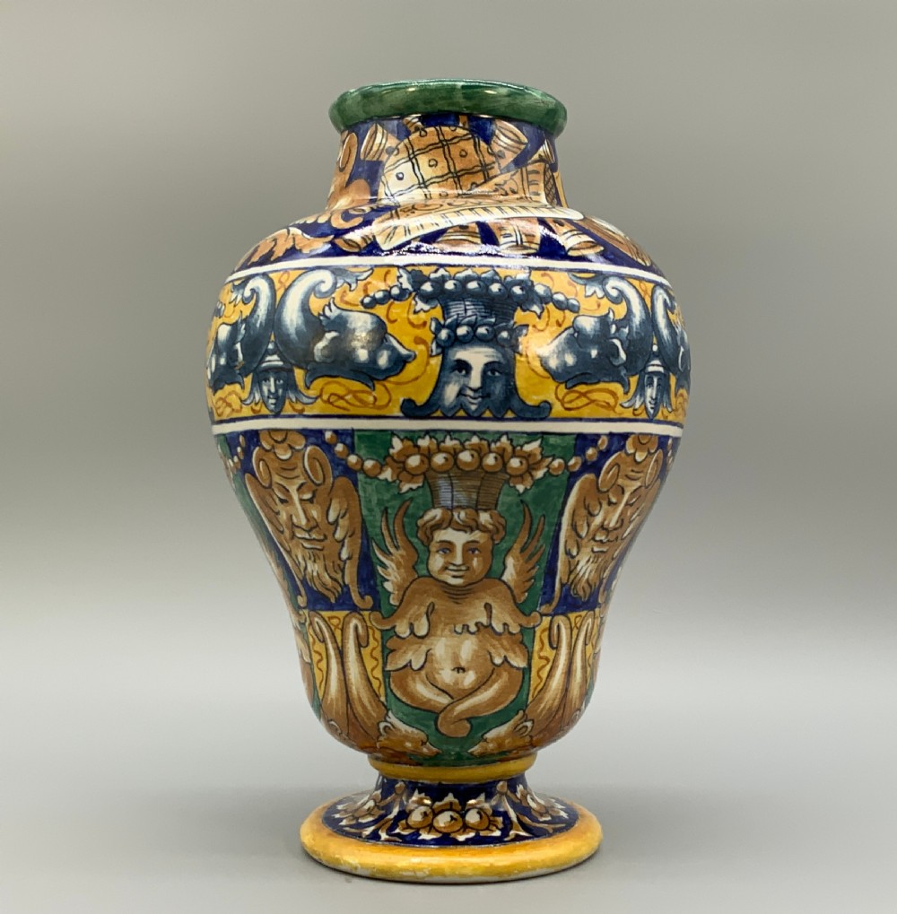 A tall hand-painted polychrome majolica ceramic vase featuring classical faces, winged cherub figures, and scrolling foliage on a circular pedestal foot.