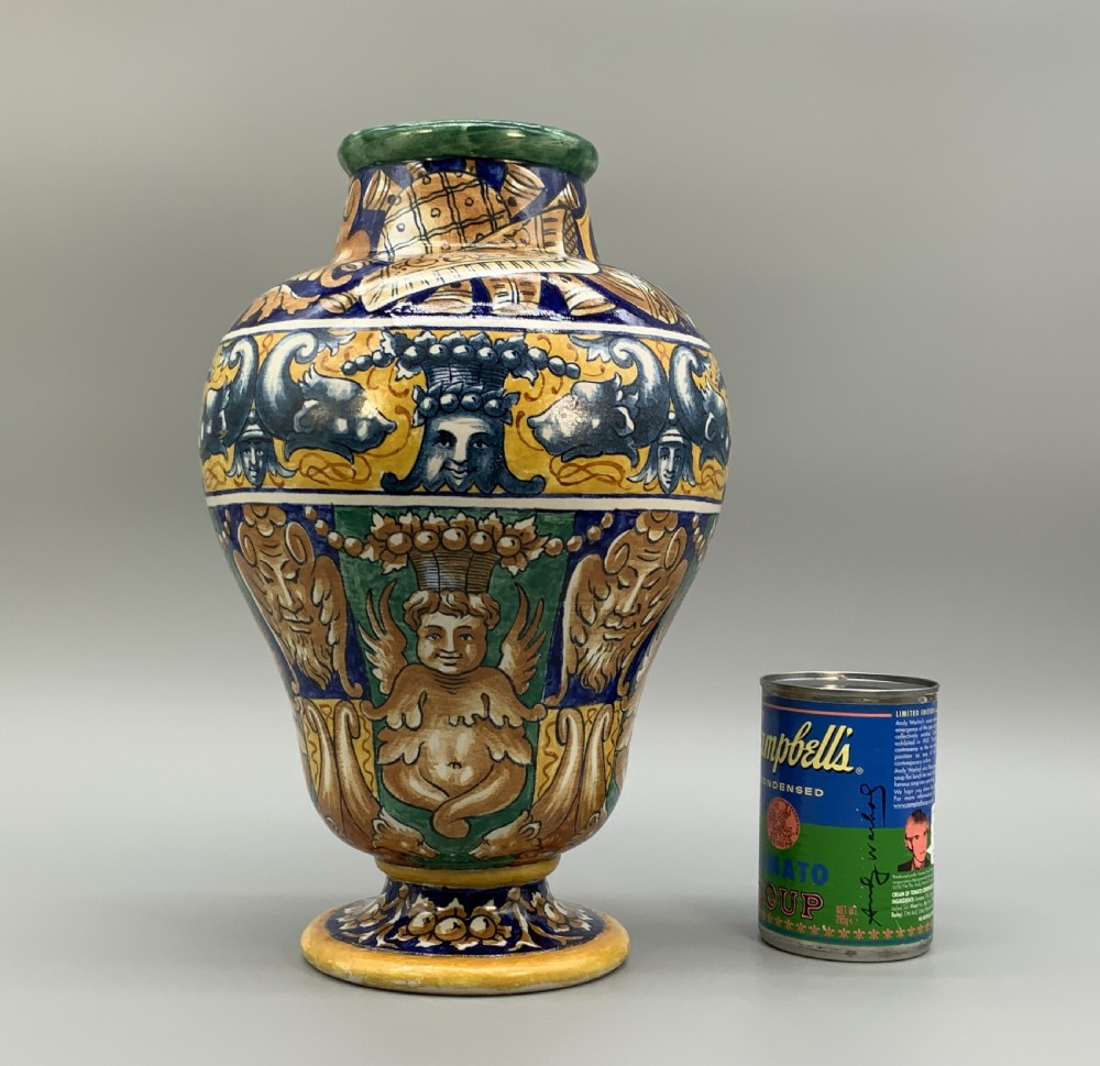 A tall hand-painted polychrome majolica ceramic vase featuring classical faces, winged cherub figures, and scrolling foliage on a circular pedestal foot. View III