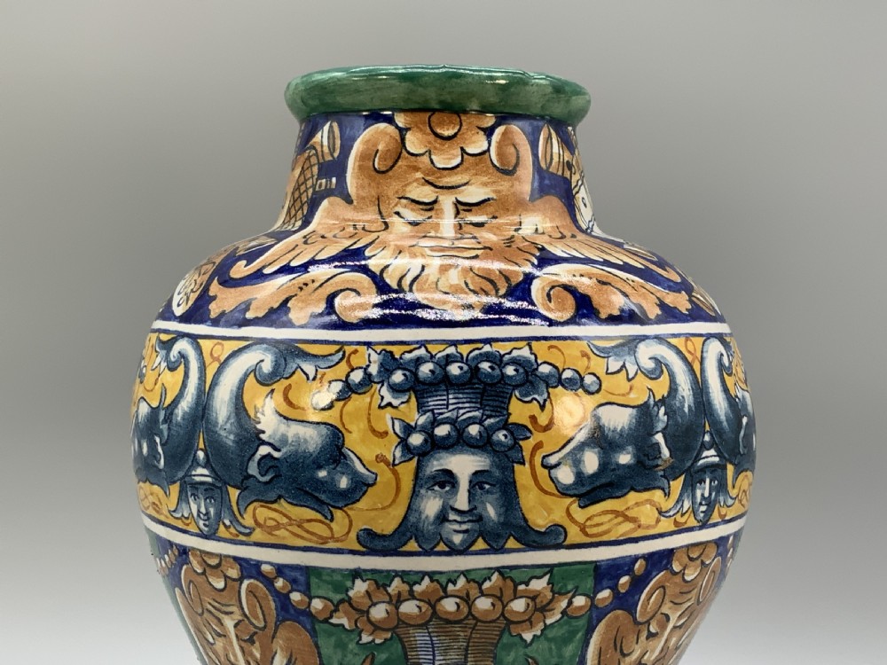 A tall hand-painted polychrome majolica ceramic vase featuring classical faces, winged cherub figures, and scrolling foliage on a circular pedestal foot. View IV