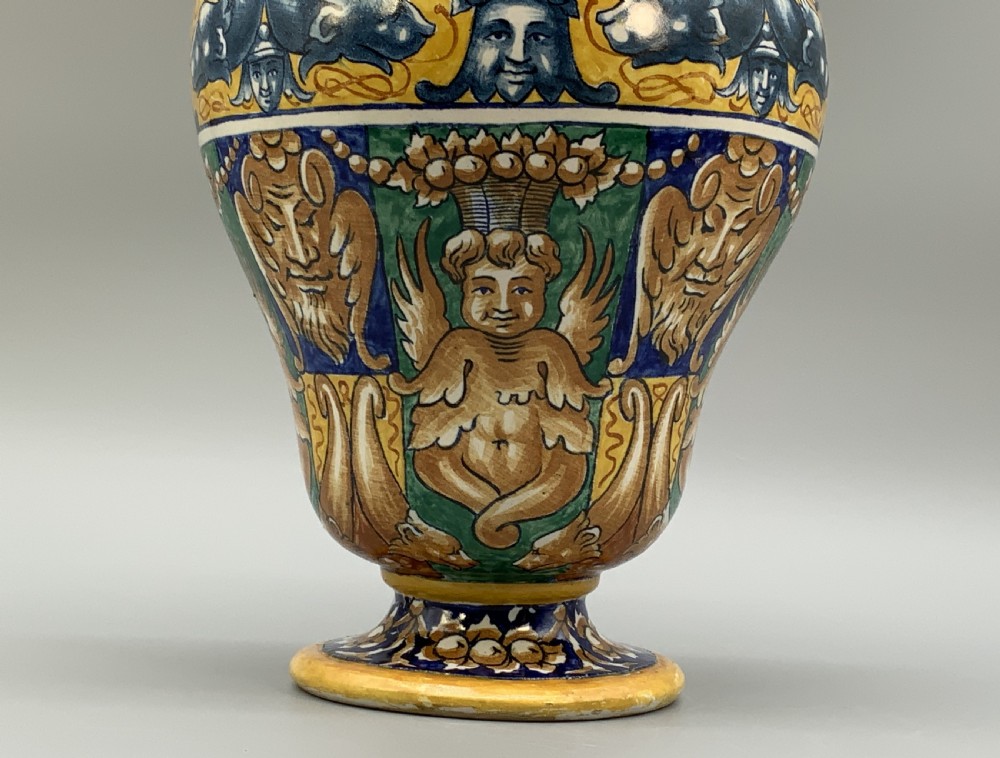 A tall hand-painted polychrome majolica ceramic vase featuring classical faces, winged cherub figures, and scrolling foliage on a circular pedestal foot. View V