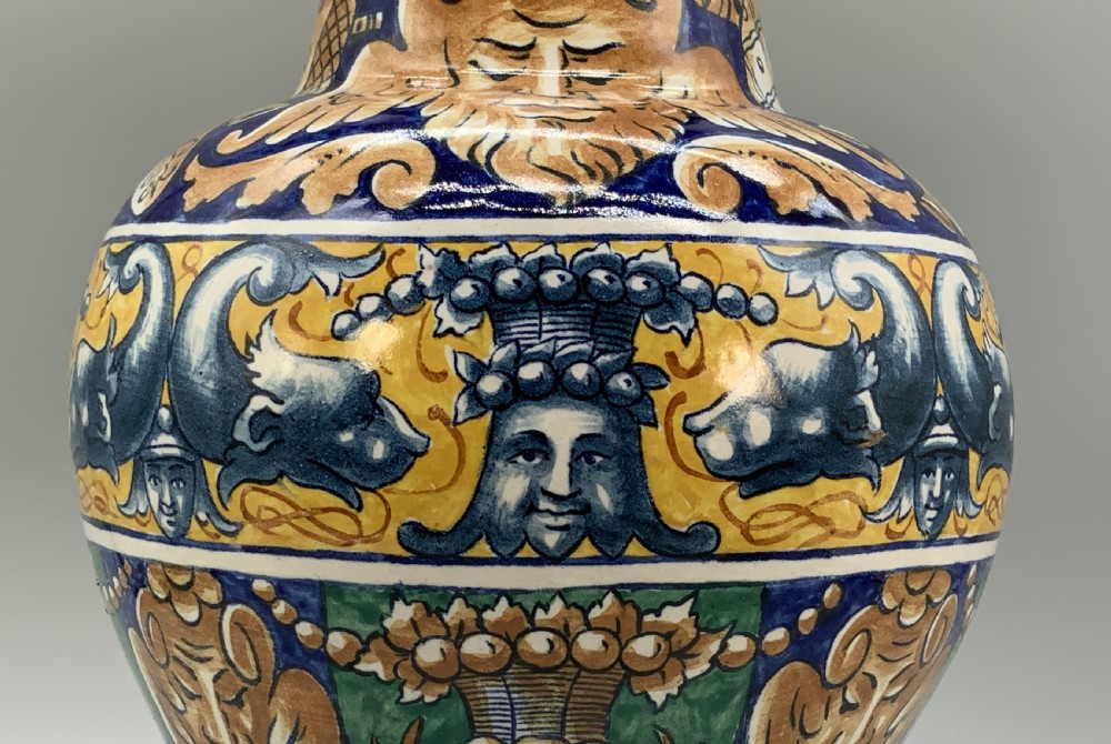A tall hand-painted polychrome majolica ceramic vase featuring classical faces, winged cherub figures, and scrolling foliage on a circular pedestal foot. View VI
