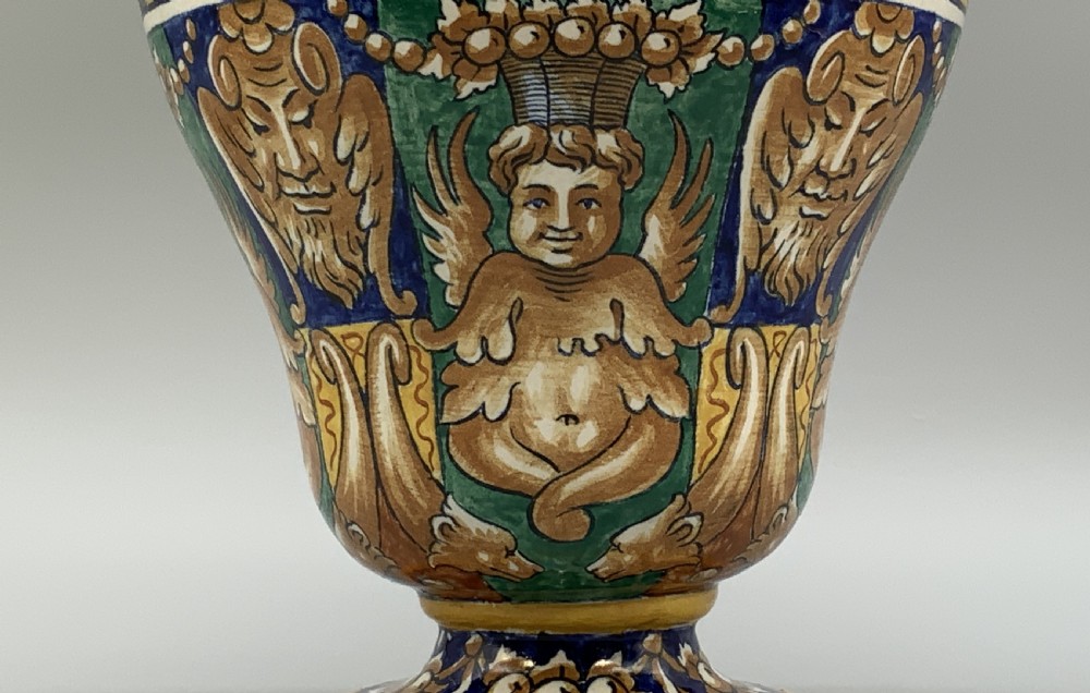A tall hand-painted polychrome majolica ceramic vase featuring classical faces, winged cherub figures, and scrolling foliage on a circular pedestal foot. View VIII