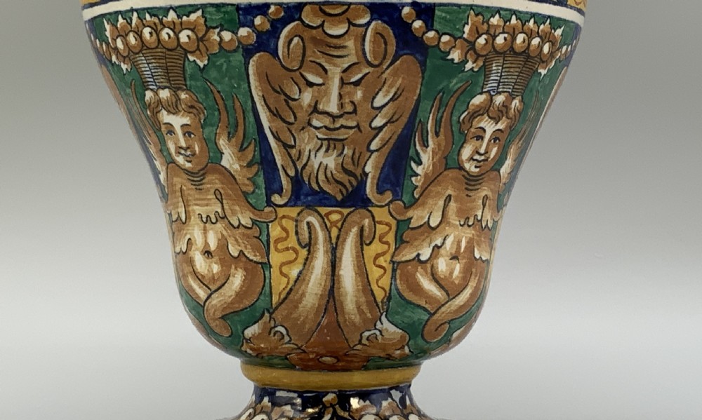 A tall hand-painted polychrome majolica ceramic vase featuring classical faces, winged cherub figures, and scrolling foliage on a circular pedestal foot. View IX