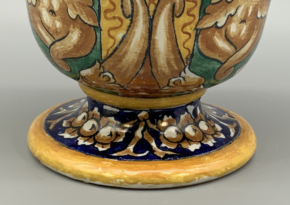 A tall hand-painted polychrome majolica ceramic vase featuring classical faces, winged cherub figures, and scrolling foliage on a circular pedestal foot. View X