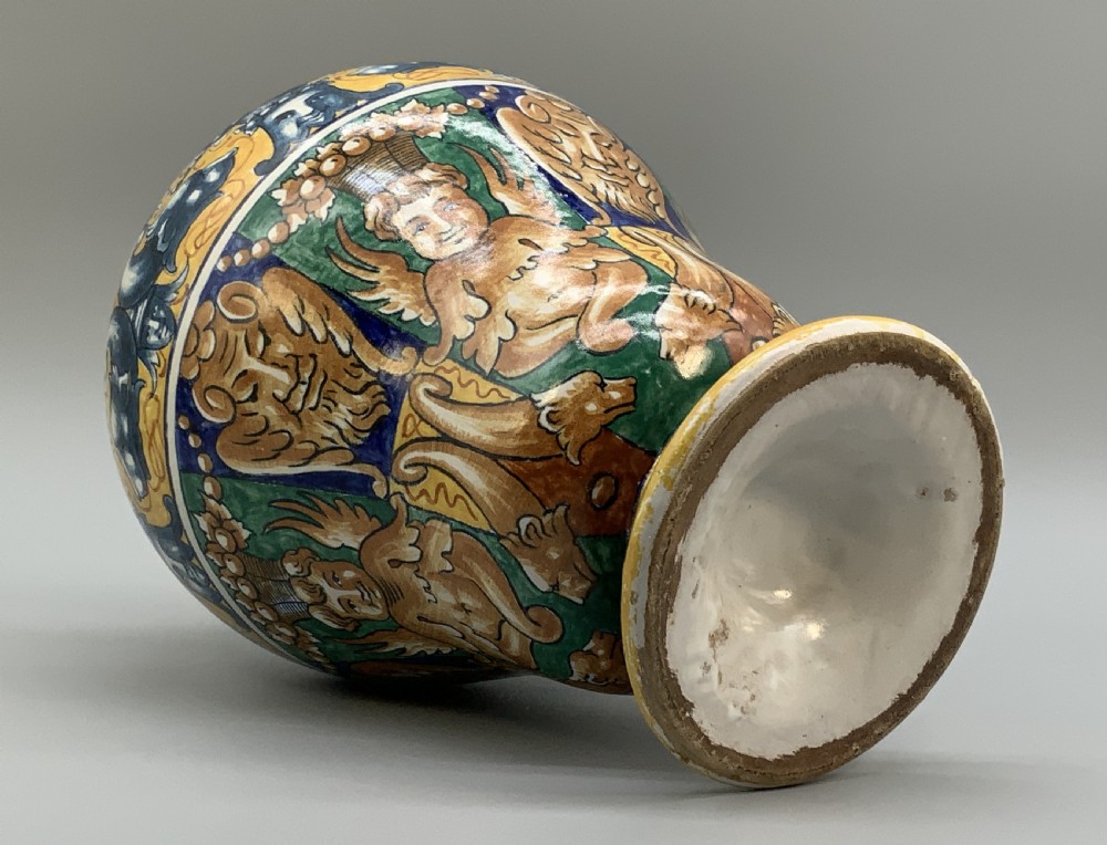 A tall hand-painted polychrome majolica ceramic vase featuring classical faces, winged cherub figures, and scrolling foliage on a circular pedestal foot. View XII