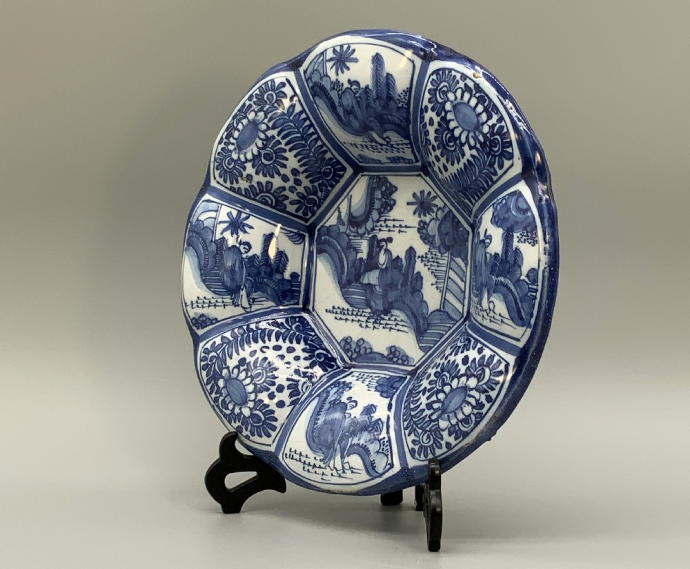 A blue and white tin-glazed earthenware lobed dish featuring panelled borders and a central Chinoiserie landscape scene. View IX
