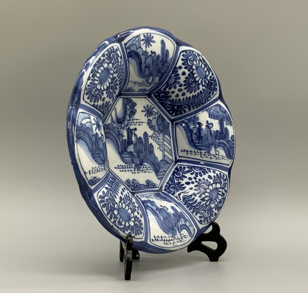 A blue and white tin-glazed earthenware lobed dish featuring panelled borders and a central Chinoiserie landscape scene. View X