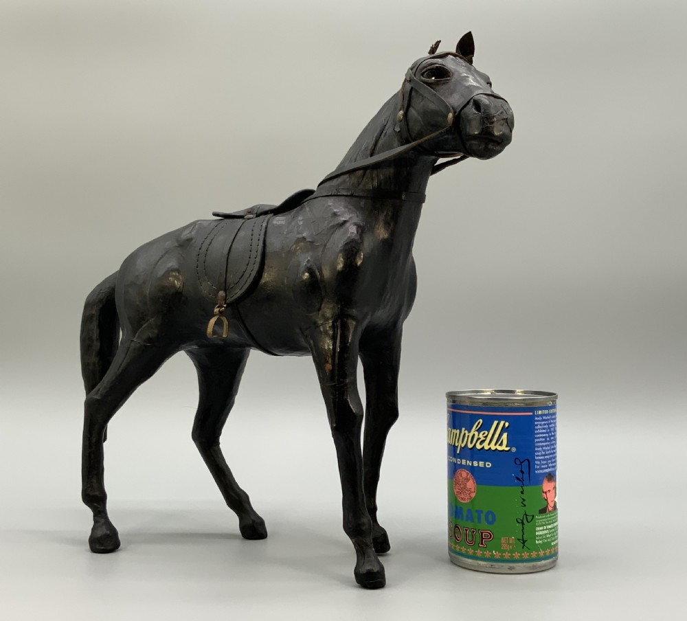 A standing vintage dark leather horse sculpture detailed with a miniature saddle, bridle, and metal stirrups. View III