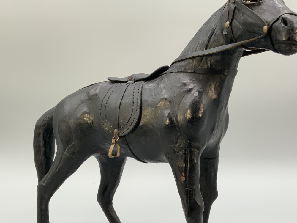 A standing vintage dark leather horse sculpture detailed with a miniature saddle, bridle, and metal stirrups. View V