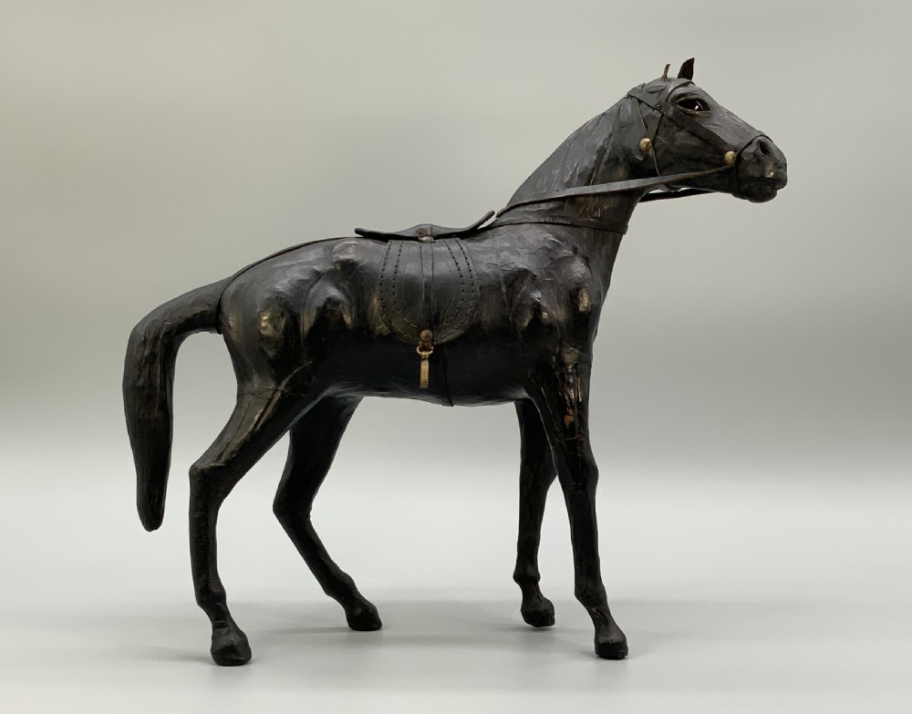A standing vintage dark leather horse sculpture detailed with a miniature saddle, bridle, and metal stirrups. View VII
