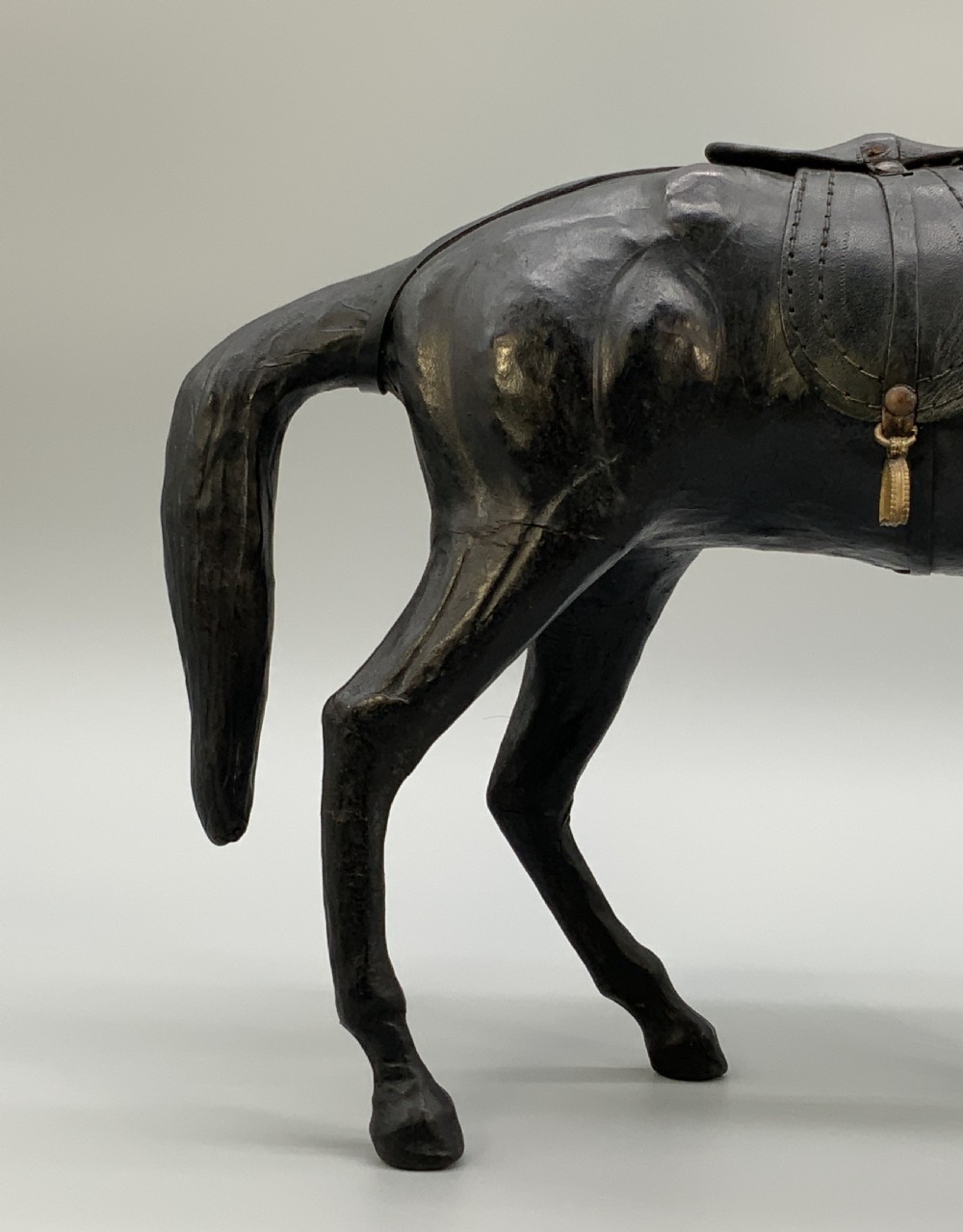 A standing vintage dark leather horse sculpture detailed with a miniature saddle, bridle, and metal stirrups. View X