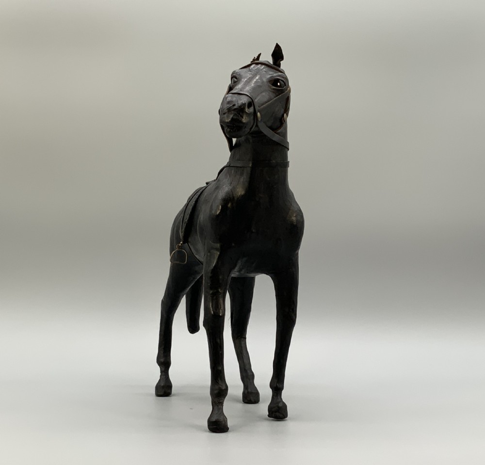 A standing vintage dark leather horse sculpture detailed with a miniature saddle, bridle, and metal stirrups. View XI