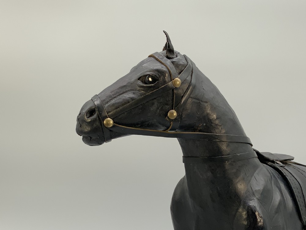 A standing vintage dark leather horse sculpture detailed with a miniature saddle, bridle, and metal stirrups. View XII