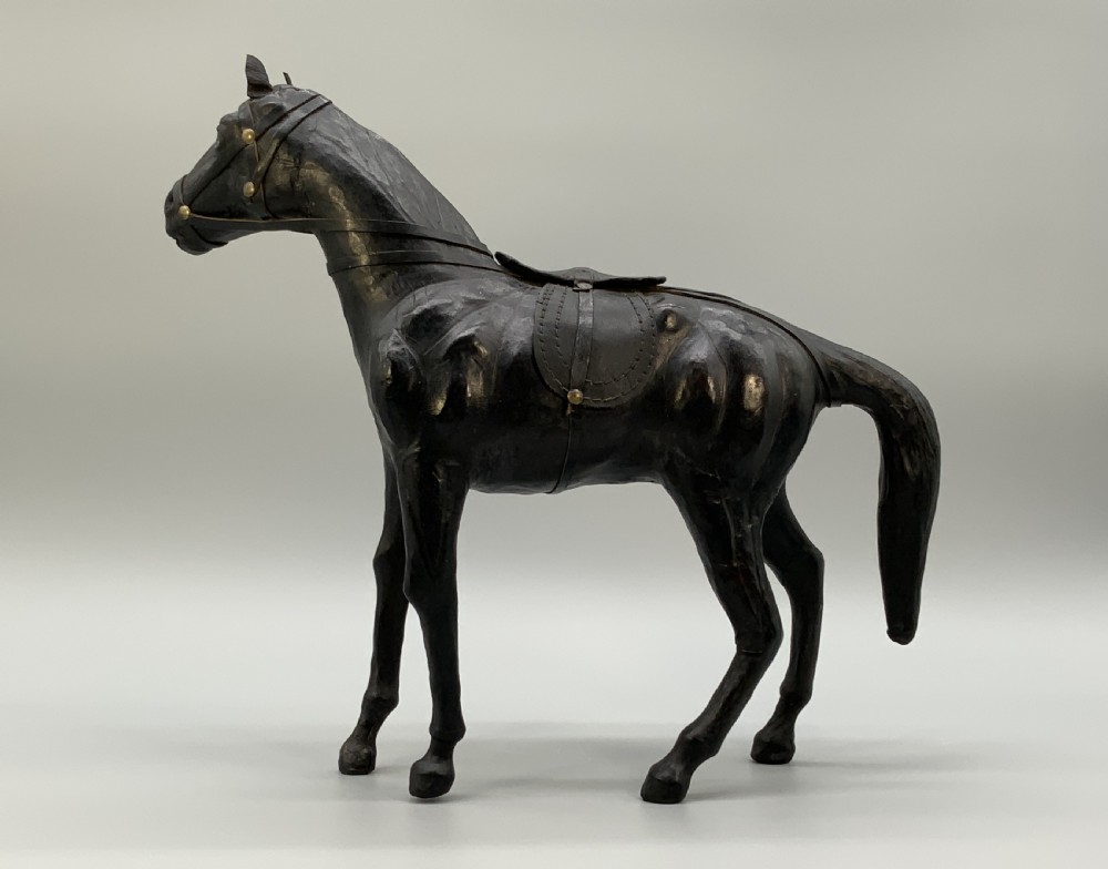 A standing vintage dark leather horse sculpture detailed with a miniature saddle, bridle, and metal stirrups. View XIII