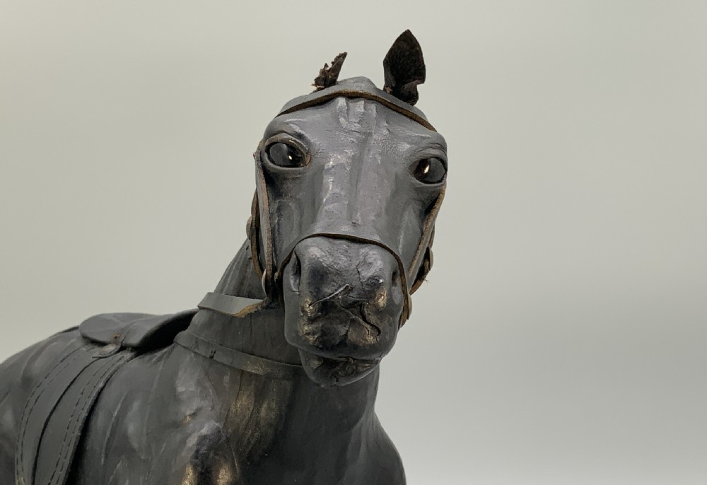 A standing vintage dark leather horse sculpture detailed with a miniature saddle, bridle, and metal stirrups. View XVI