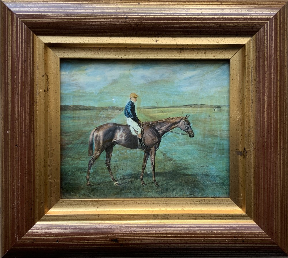 A framed miniature print depicting a jockey in blue silks seated on a dark racehorse in a countryside landscape, enclosed in a moulded timber frame with a gold inner slip. View II