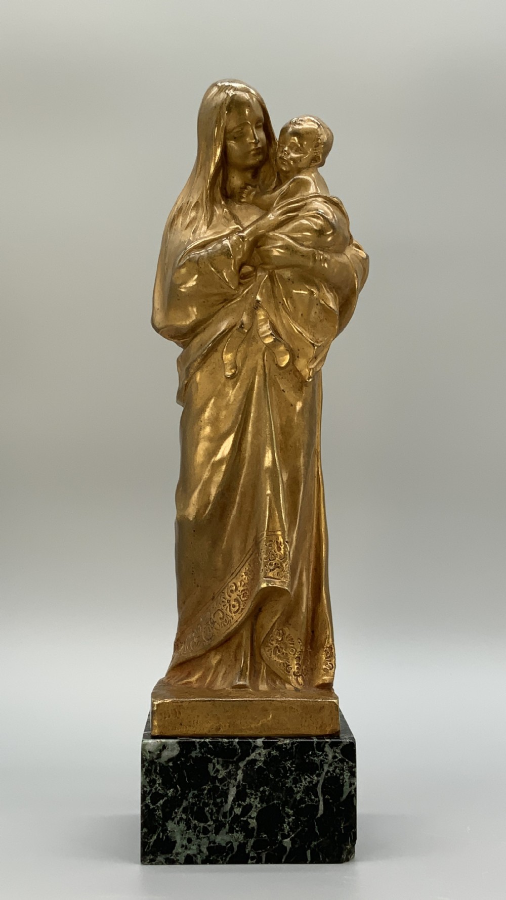 A gilt bronze sculpture depicting the Madonna holding the infant Christ, mounted on a square dark green marbled base.