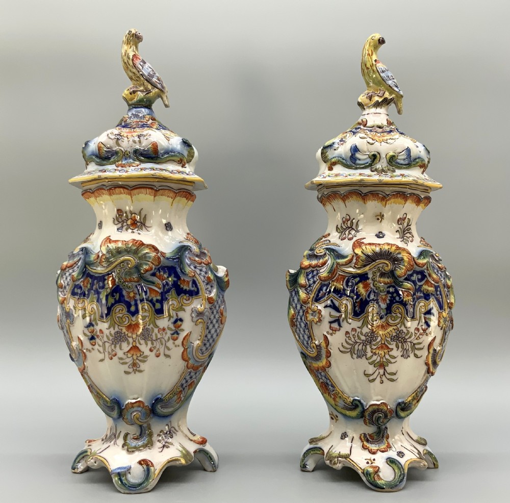 A pair of ornate French faience lidded vases decorated with hand-painted polychrome florals, Rococo scrollwork moulding, and sculptural parrot finials.