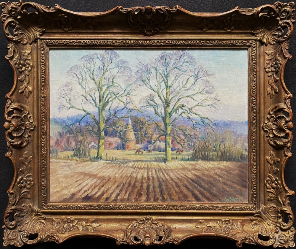 An original landscape oil painting depicting a ploughed field and bare trees before a hop kiln, set in an ornate swept gilded frame.
