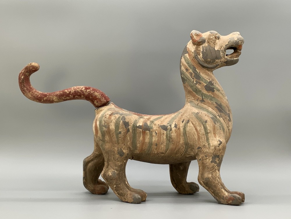 A painted earthenware figure of a tiger-like mythical beast from the Han Dynasty featuring an arched neck, raised tail, and red and green painted stripes.