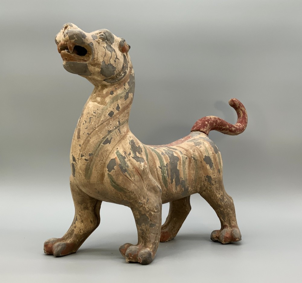 A painted earthenware figure of a tiger-like mythical beast from the Han Dynasty featuring an arched neck, raised tail, and red and green painted stripes. View IV