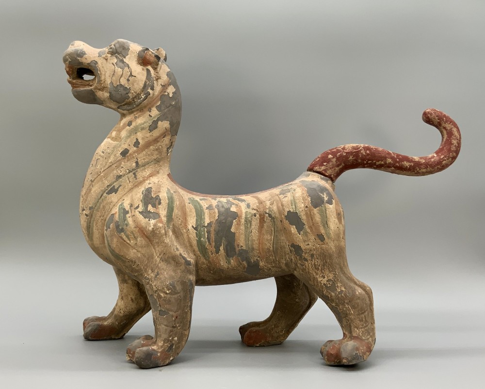 A painted earthenware figure of a tiger-like mythical beast from the Han Dynasty featuring an arched neck, raised tail, and red and green painted stripes. View V