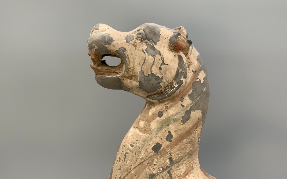 A painted earthenware figure of a tiger-like mythical beast from the Han Dynasty featuring an arched neck, raised tail, and red and green painted stripes. View VI