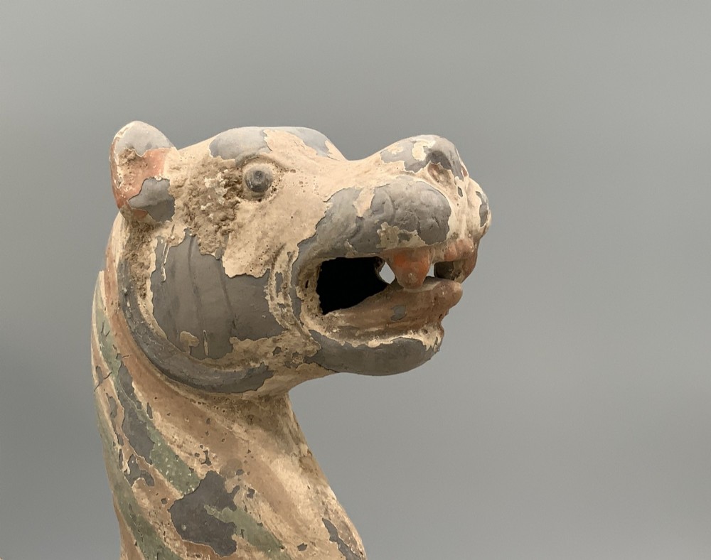 A painted earthenware figure of a tiger-like mythical beast from the Han Dynasty featuring an arched neck, raised tail, and red and green painted stripes. View IX
