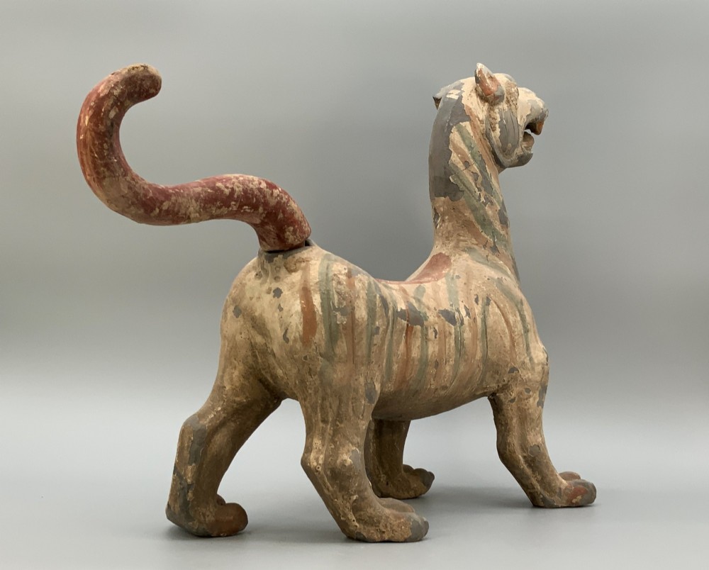 A painted earthenware figure of a tiger-like mythical beast from the Han Dynasty featuring an arched neck, raised tail, and red and green painted stripes. View X