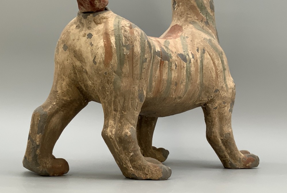A painted earthenware figure of a tiger-like mythical beast from the Han Dynasty featuring an arched neck, raised tail, and red and green painted stripes. View XI