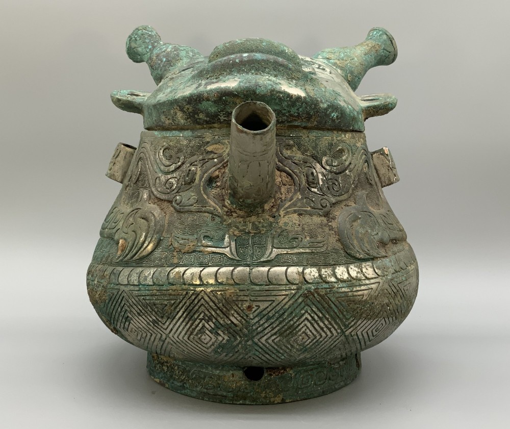 A cast bronze ritual vessel featuring a stylised human face with horn-like projections, a small tubular spout, and textured surface oxidisation. View V