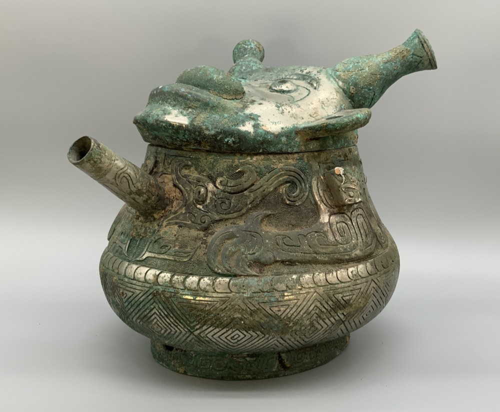 A cast bronze ritual vessel featuring a stylised human face with horn-like projections, a small tubular spout, and textured surface oxidisation. View VI
