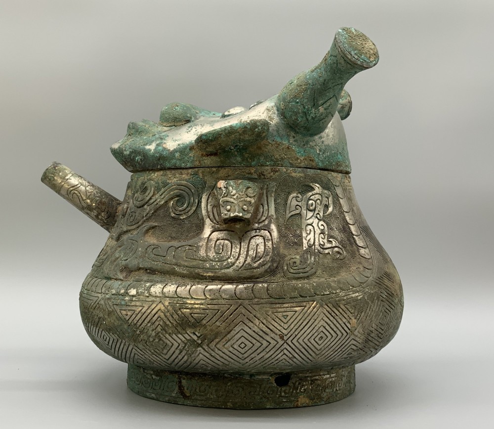 A cast bronze ritual vessel featuring a stylised human face with horn-like projections, a small tubular spout, and textured surface oxidisation. View VII