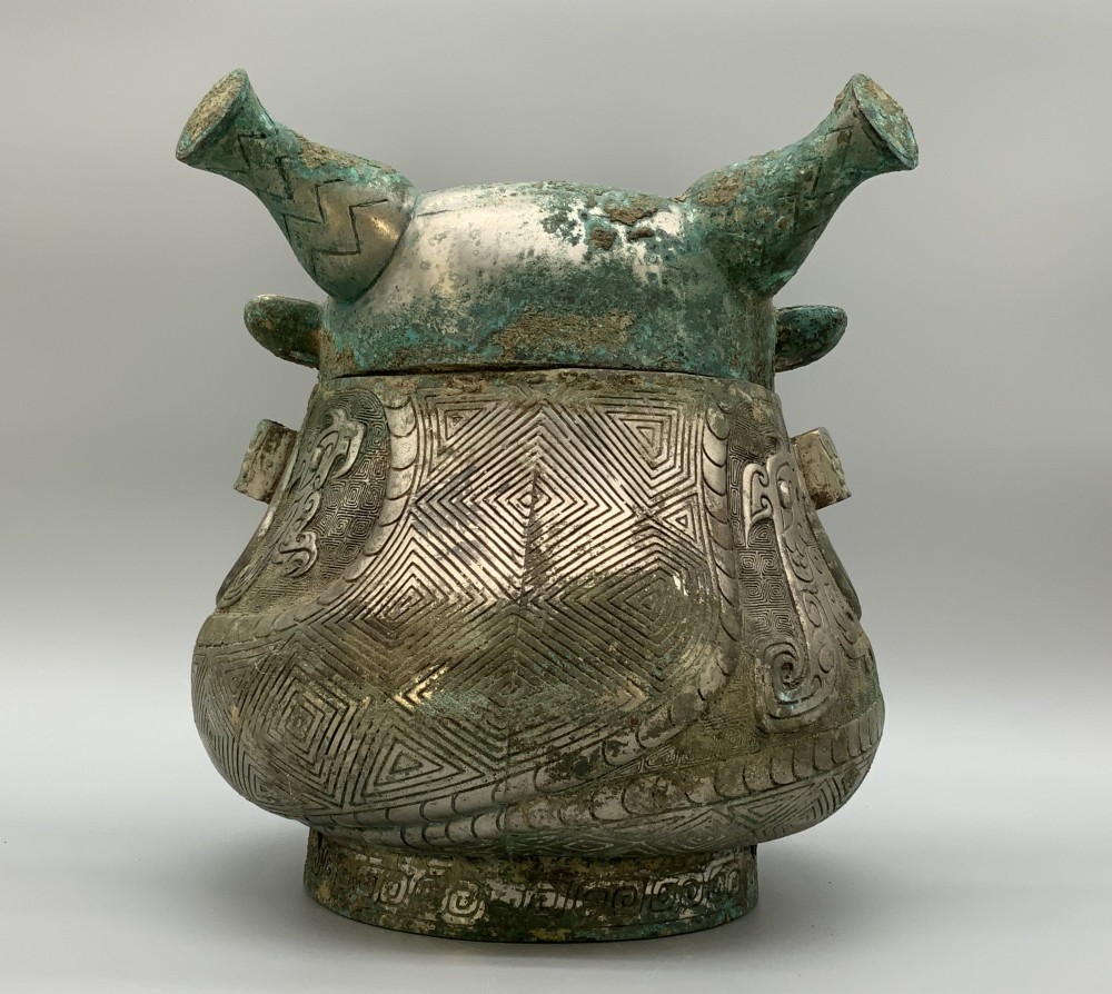 A cast bronze ritual vessel featuring a stylised human face with horn-like projections, a small tubular spout, and textured surface oxidisation. View VIII
