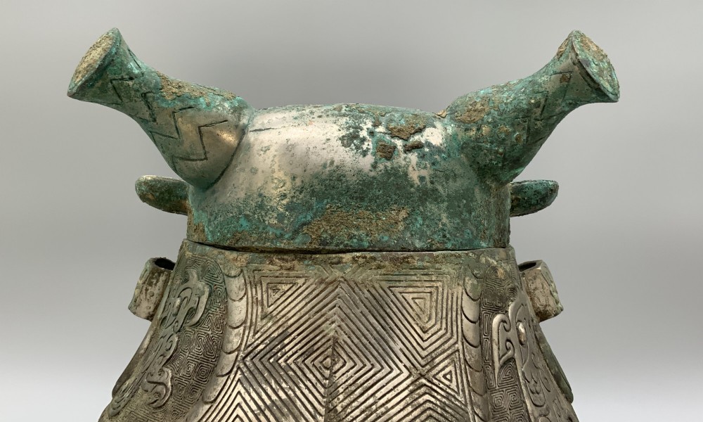 A cast bronze ritual vessel featuring a stylised human face with horn-like projections, a small tubular spout, and textured surface oxidisation. View IX