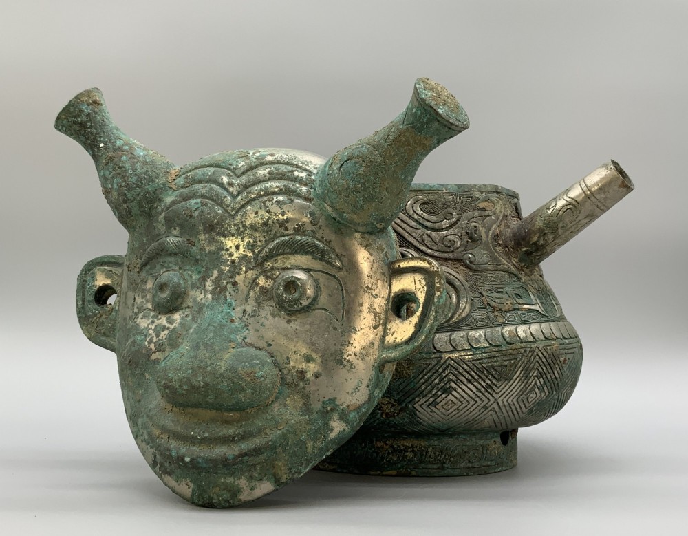 A cast bronze ritual vessel featuring a stylised human face with horn-like projections, a small tubular spout, and textured surface oxidisation. View X