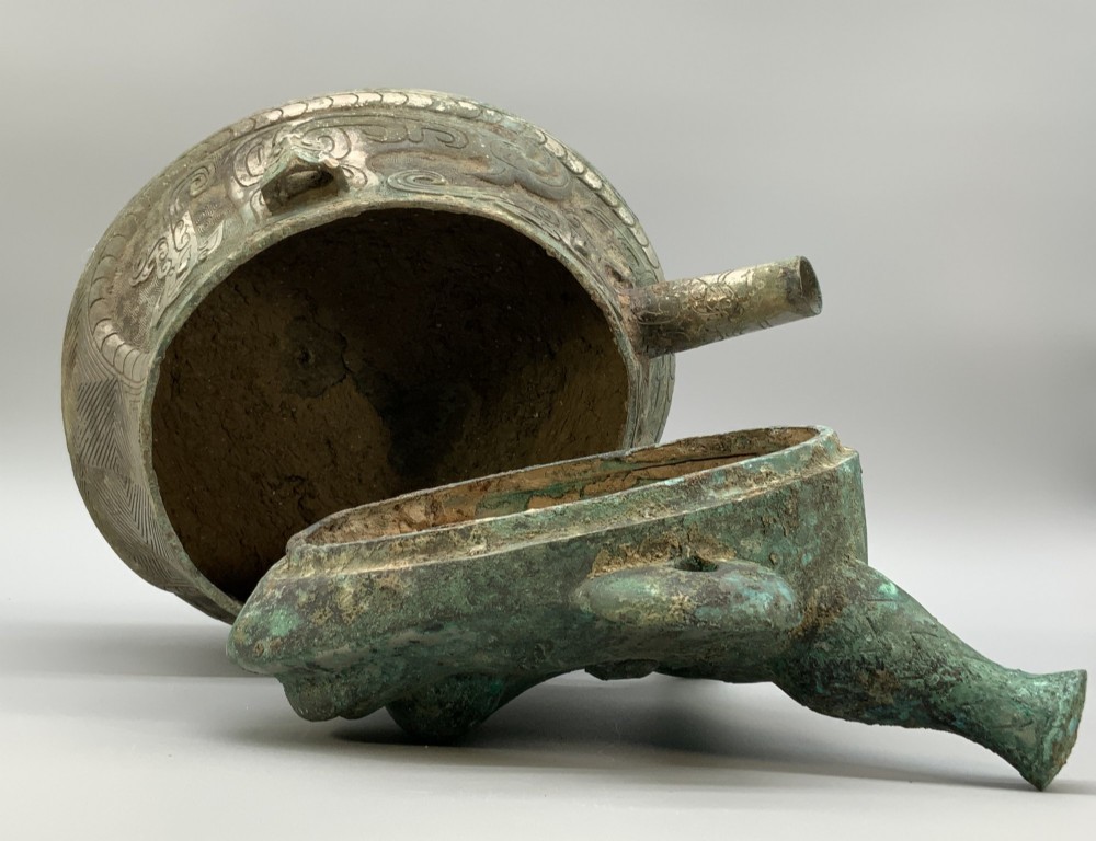 A cast bronze ritual vessel featuring a stylised human face with horn-like projections, a small tubular spout, and textured surface oxidisation. View XI