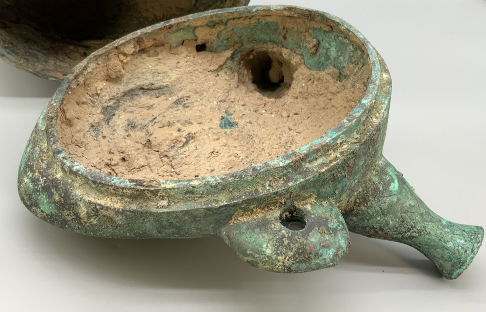 A cast bronze ritual vessel featuring a stylised human face with horn-like projections, a small tubular spout, and textured surface oxidisation. View XII