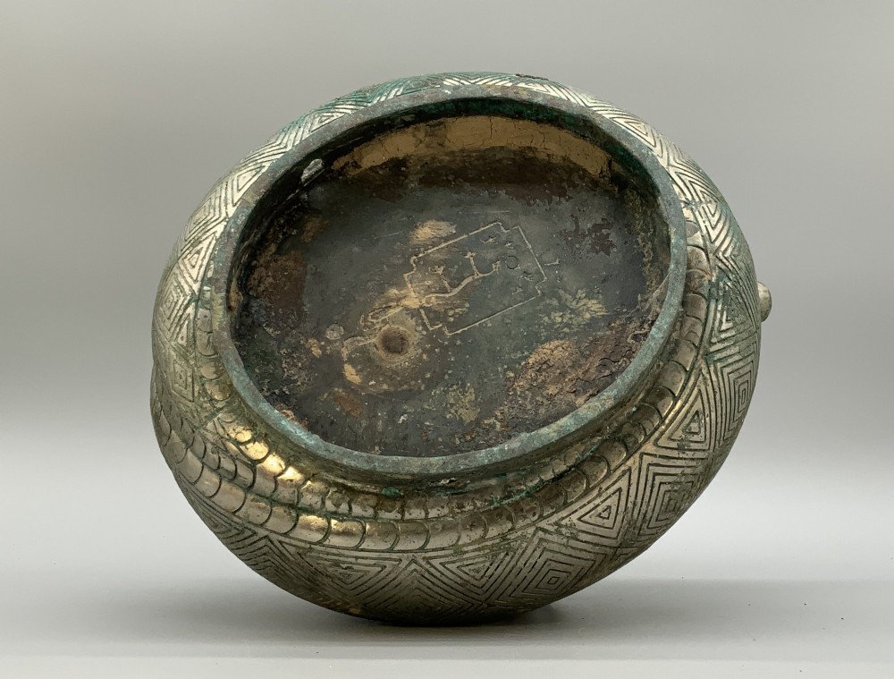 A cast bronze ritual vessel featuring a stylised human face with horn-like projections, a small tubular spout, and textured surface oxidisation. View XIII