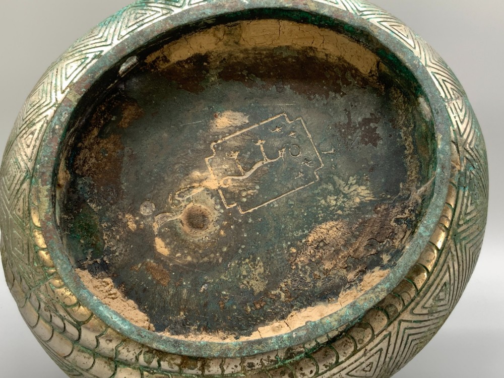 A cast bronze ritual vessel featuring a stylised human face with horn-like projections, a small tubular spout, and textured surface oxidisation. View XIV