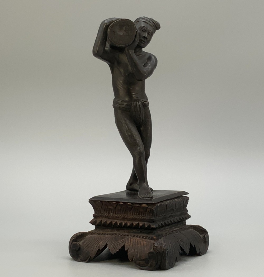 A dark patinated bronze figurine of a man carrying a log on his shoulder, mounted on an intricately carved wooden stand. View V