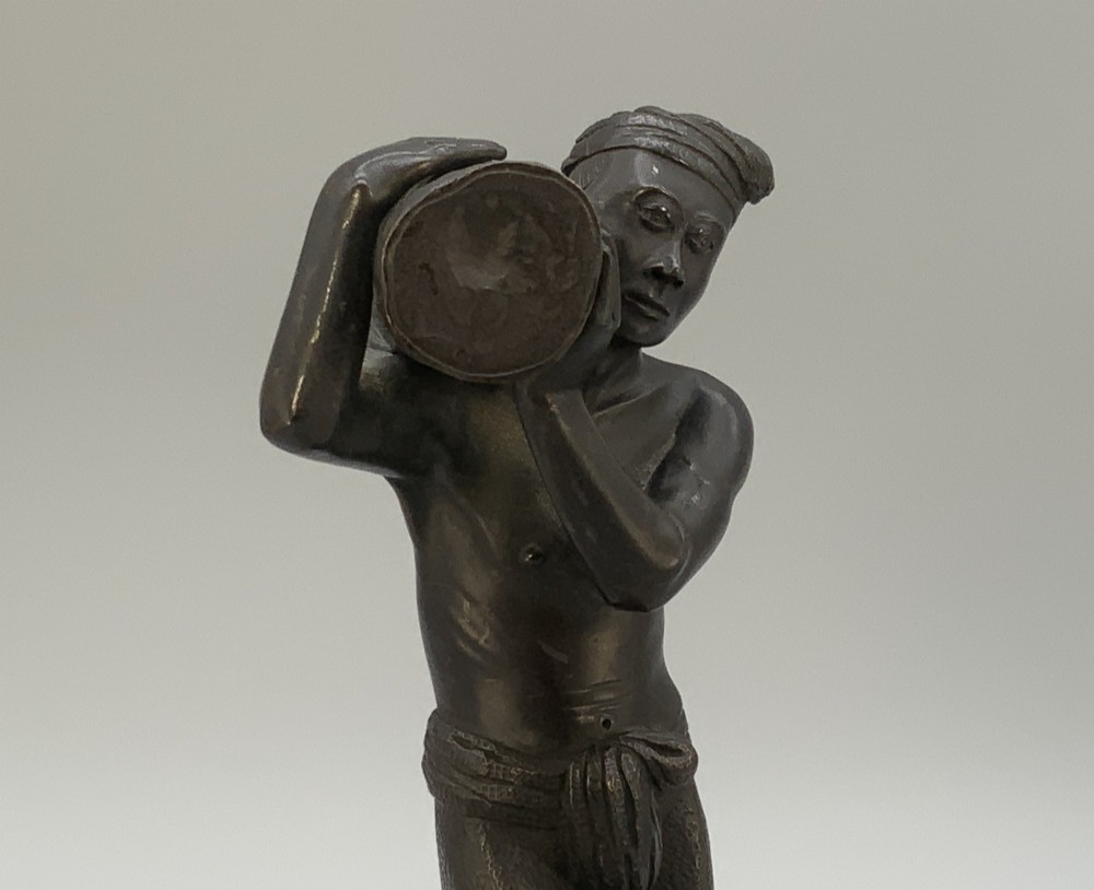 A dark patinated bronze figurine of a man carrying a log on his shoulder, mounted on an intricately carved wooden stand. View VI