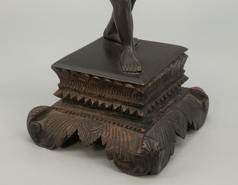A dark patinated bronze figurine of a man carrying a log on his shoulder, mounted on an intricately carved wooden stand. View VII